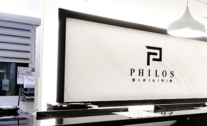 접수대 뒤 흰 대리석 벽의 PHILOS 로고 간판과 펜던트 조명